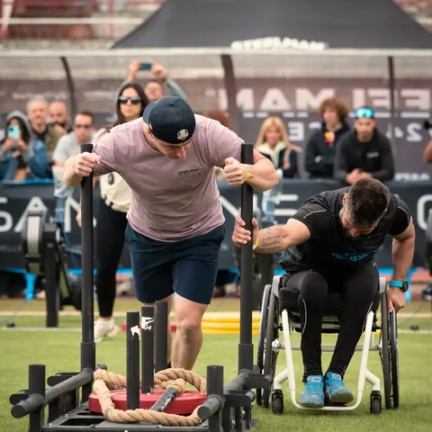 Two athletes pushing weighted sleds across grass, one of them a wheelchair user, with spectators behind.